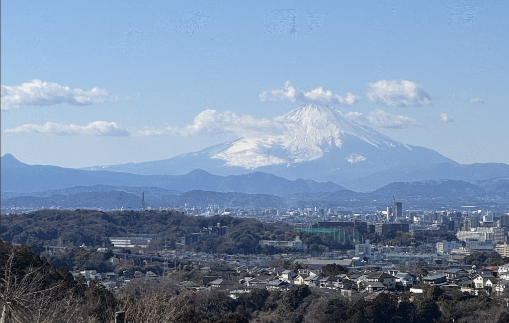 A distant Mt. Fuji with snow covering most of its flank soars above a Japanese cityscape and wooded hills.